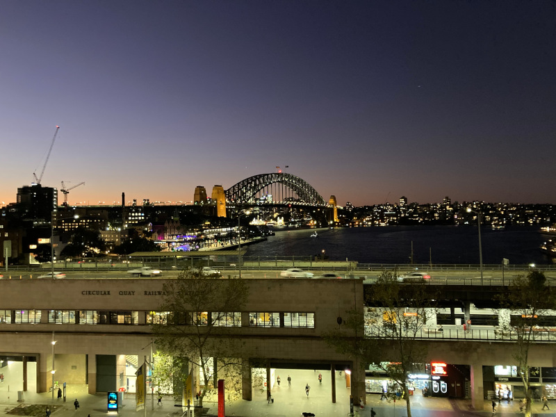 A cityscape at dusk featuring the Sydney Harbour Bridge and the waterfront, with illuminated buildings and a railway station in the foreground. The sky transitions from orange to deep blue, with a few clouds and city lights visible.